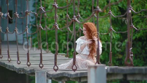 迷人的可爱的年轻女子一头火红的头发坐在河上的吊桥上梳着辫子视频的预览图