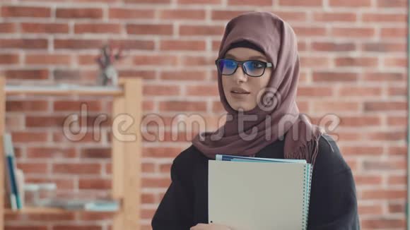 一位带着文件的穆斯林商务女士看着相机微笑一个戴着头巾的年轻女孩站在办公室里和一个视频的预览图