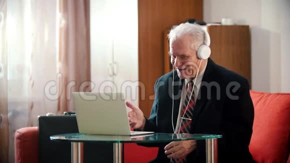 年迈的祖父戴着耳机的老祖父微笑着看着房间里的笔记本电脑屏幕视频的预览图