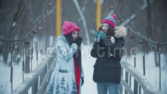 两个年轻的女朋友站在雪桥上喝着热水瓶里的热饮料视频的预览图