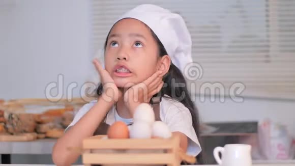一个戴着白色厨师帽的亚洲小女孩正坐在厨房的木篮里玩鸡蛋视频的预览图