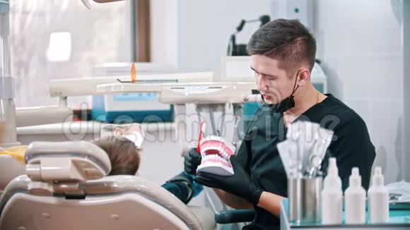 一位年轻的牙医向一个小男孩展示了如何在人体下巴的塑料模型上刷牙视频的预览图