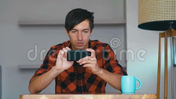 年轻人拿着智能手机玩手机游戏坐在家里的桌子旁快乐的年轻人使用社交媒体应用程序工作学习视频的预览图