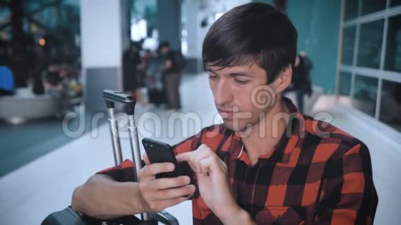 年轻人穿着格子衬衫带着行李在机场候机楼使用智能手机等待登机视频的预览图