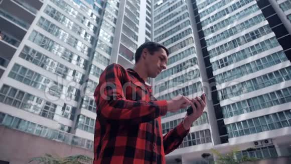 英俊的年轻人穿着格子衬衫在城市建筑背景下浏览他的智能手机视频的预览图