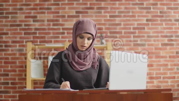 一个穆斯林女孩用钢笔写信息并在她的笔记本电脑上检查一个戴头巾的年轻女孩正在办公室工作视频的预览图