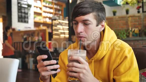 现代咖啡馆里使用智能手机触摸屏的人男性饮用奶昔或冰沙并在智能手机上发短信视频的预览图