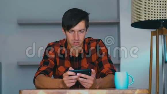 年轻人拿着智能手机玩手机游戏坐在家里的桌子旁快乐的年轻人使用社交媒体应用程序工作学习视频的预览图