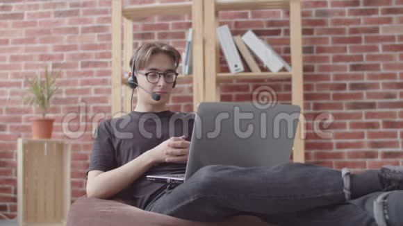 一位年轻的接线员正在通过笔记本电脑上的麦克风通过视频聊天交谈一个年轻人坐在包椅上咨询视频的预览图