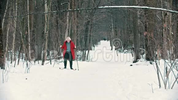一个穿着红色羽绒服的年轻金发女人在森林里滑雪时正要摔倒视频的预览图
