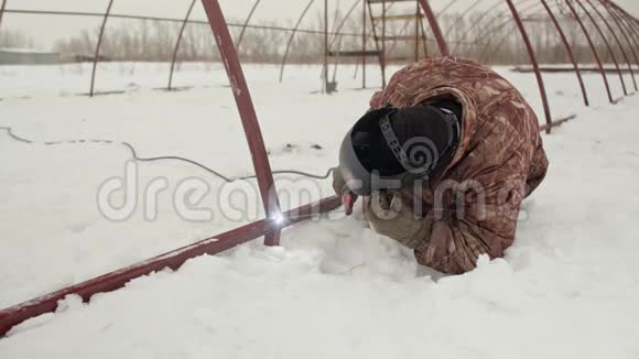 一个穿着温暖的冬装和焊接面罩的人正在地面上焊接一个金属结构专业焊工焊接视频的预览图