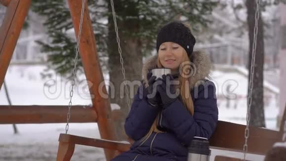 下雪时年轻女子坐在长凳上喝热饮料视频的预览图