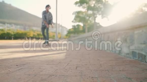 阳光明媚的一天一个年轻人骑在滑板上朝坐在公园墙上的摄像机迈开双脚视频的预览图