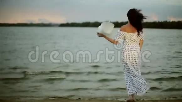 一位穿着夏日长裙的中年妇女站在海滩上脱下帽子享受着风视频的预览图