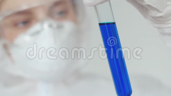 一个戴着医用口罩和安全眼镜的实验室助理女孩拿着一瓶蓝色溶液在她面前看着它视频的预览图