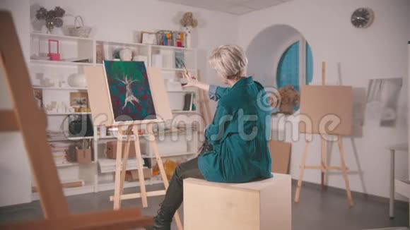 一位金发短发的年轻女子坐在艺术工作室里远远地看着她的画视频的预览图