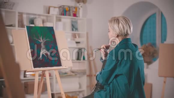 一位金发短发的年轻女艺术家坐在艺术工作室里从远处看着她的画视频的预览图