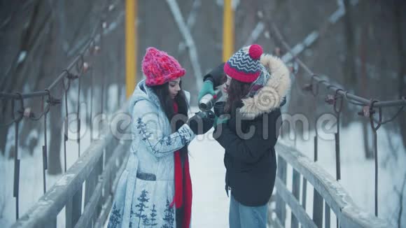 两个年轻的女人站在雪桥上喝着热水瓶里的热饮料视频的预览图
