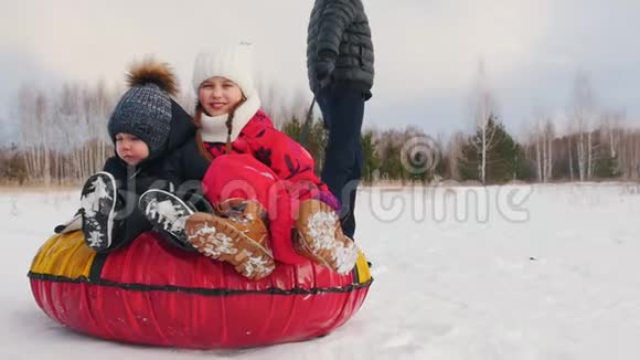 一家人在冬天的森林附近玩耍父亲骑着他的孩子在充气雪橇上玩耍视频的预览图