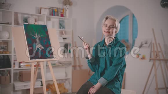 一位年轻的女艺术家拿着画笔坐在艺术工作室的椅子上视频的预览图