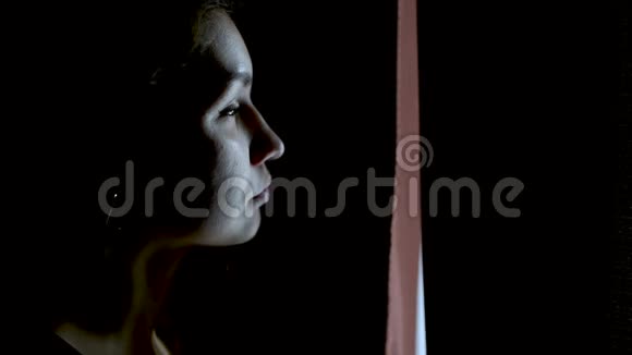 一个在黑暗的房间里悲伤的女孩打开一扇窗帘望着窗外视频的预览图