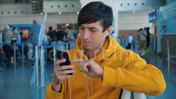 年轻的男性穿着黄色连帽衫在机场等候飞机使用带耳机的智能手机视频的预览图