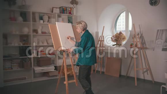 一位年轻的金发女艺术家在画布上画素描在耳机里听音乐视频的预览图