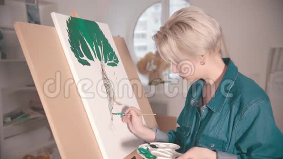一个年轻的金发女人在艺术工作室画一棵画着颜料的树视频的预览图