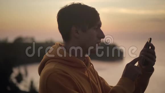 在美丽的日落背景下男性旅行者的剪影在海洋上方的山上使用智能手机视频的预览图