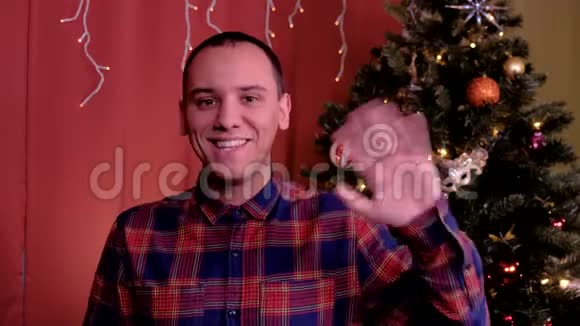 一个男人看着相机挥手问候圣诞节背景中的圣诞树假期概念视频的预览图