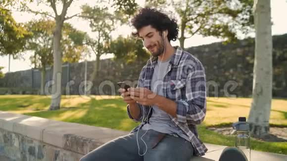 一个英俊的微笑的年轻人坐在公园里的肖像他的水瓶和耳机在他的耳朵发短信视频的预览图