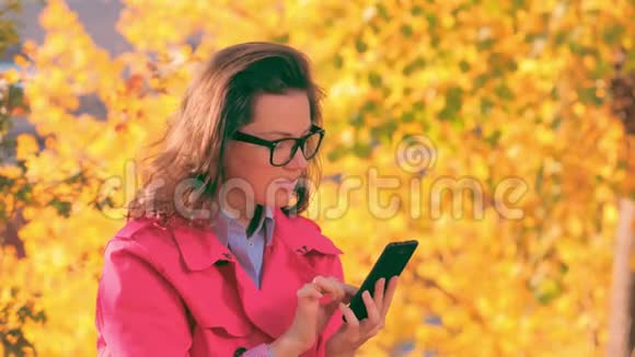 在秋天公园一个穿着红色夹克的女人在智能手机上发短信视频的预览图