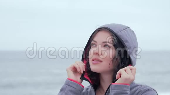 一个穿着夹克带着兜帽的蓝眼睛女人在海滩上的肖像视频的预览图