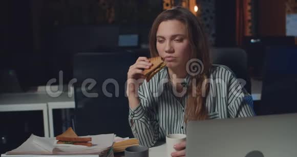 沮丧的女办公室经理在夜间办公室吃三明治和喝咖啡精疲力竭的女孩吃晚饭视频的预览图