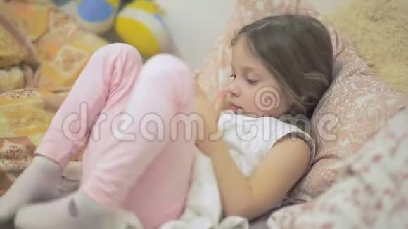 一个小女孩躺在床上在智能手机屏幕上看动画片儿童和数字技术视频的预览图
