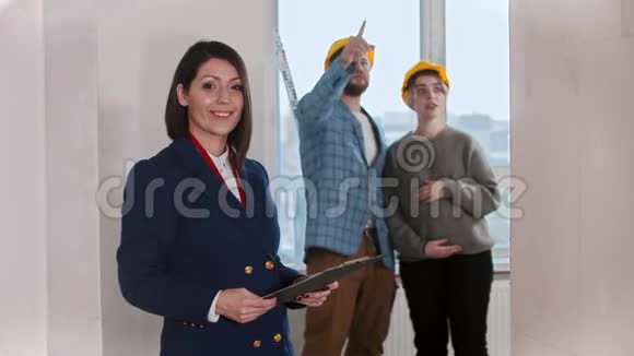 一位房地产经纪人站在公寓里和一对微笑的年轻夫妇在后台环顾四周视频的预览图