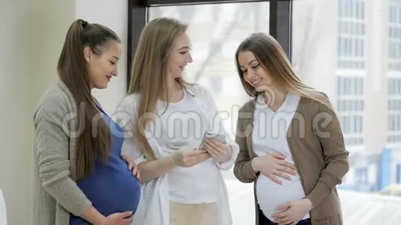 一群快乐的孕妇站在窗户旁边观看背景和使用手机视频的预览图
