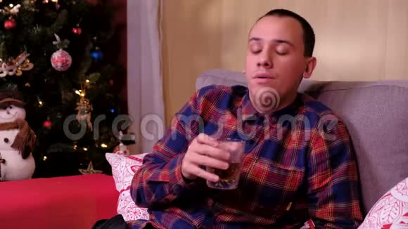 一个孤独的人坐在圣诞树附近的沙发上喝威士忌的肖像假期和新年的概念视频的预览图