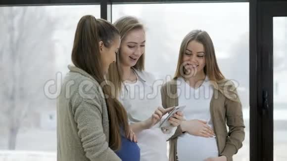 一群快乐的孕妇站在窗户旁边观看背景和使用手机视频的预览图