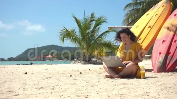 在热带沙滩上穿黄色衣服的女孩在皮划艇附近的笔记本电脑上工作喝新鲜芒果远程工作成功视频的预览图