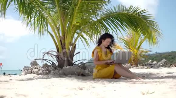 在热带沙滩上穿黄色衣服的女孩在棕榈树附近的笔记本电脑上工作喝新鲜芒果远程工作成功视频的预览图