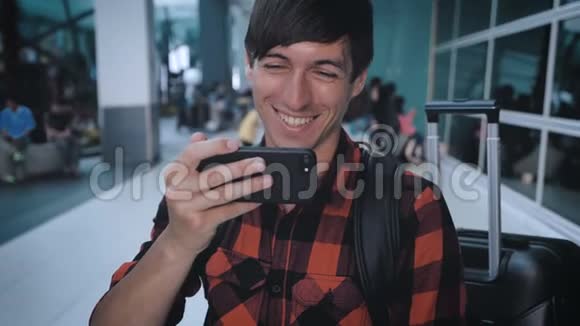 微笑男游客穿着格子衬衫带着行李在机场候机楼观看智能手机视频等待视频的预览图