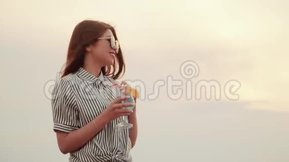 戴着太阳镜的亚洲女孩拿着一杯明亮的酒精鸡尾酒跳舞慢动作夏季城市鸡尾酒视频的预览图