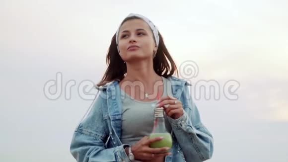 一个手里拿着绿色鸡尾酒的漂亮女孩站在户外享受着美丽的景色慢动作夏天视频的预览图