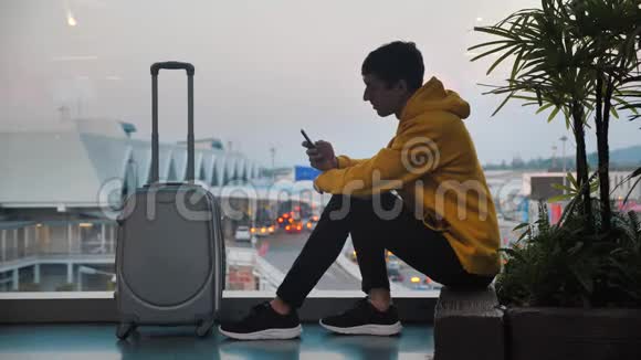 年轻的微笑男游客带着行李在机场候机楼等候窗户整洁旅客发短信视频的预览图