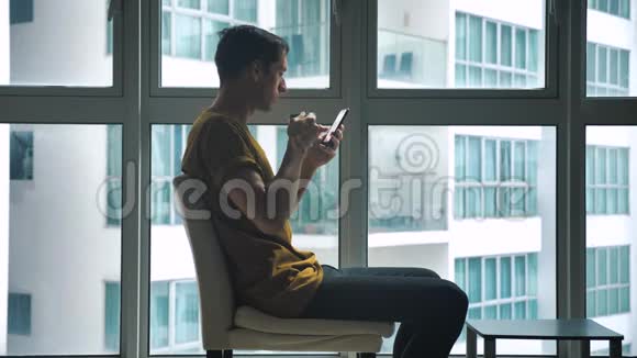 年轻人坐在窗前早上在家喝咖啡浏览智能手机视频的预览图