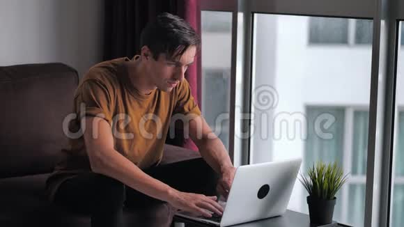 英俊的年轻人在家坐在窗前的沙发上用笔记本电脑视频的预览图