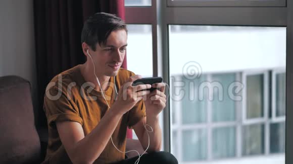戴着耳机的快乐男人喜欢在社交网络上观看媒体的智能手机而坐在家里的沙发上视频的预览图