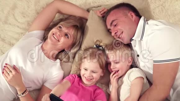 妈妈爸爸女儿和儿子在柔软的毯子上休息看着装着小女儿的平板电脑快乐的人视频的预览图