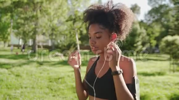 快乐运动员非裔美国女跑步者把耳机放在耳朵里在袖标里听智能手机上的音乐视频的预览图